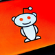 Reddit's smiling Alien logo, named Snoo, is shown against an orange backdrop on a phone.